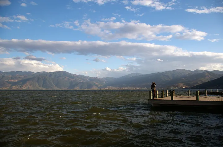 A broad blue lake with chop on the water seen from the shore, a timber-and-chain viewing platform jutting in at the right with one person standing on it, and a long dark mountain ridge running the full width of the far shore under cumulus cloud.