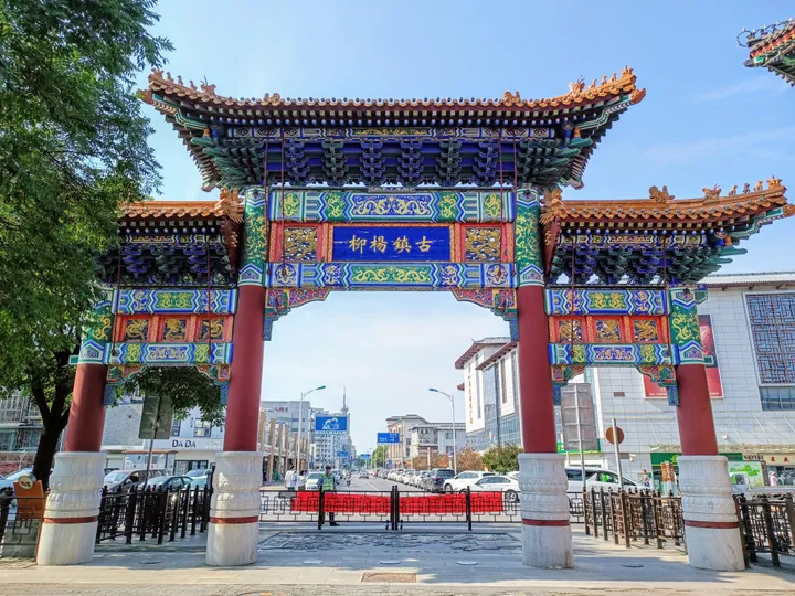The 古镇杨柳 entrance paifang standing across a street: four red piers on white stone drums carrying painted blue-and-green beams and a blue plaque lettered 古镇杨柳 in gold, with glazed-tile roofs and ridge figures; a red barrier, railings and shoppers on the paving below and shopfronts beyond.