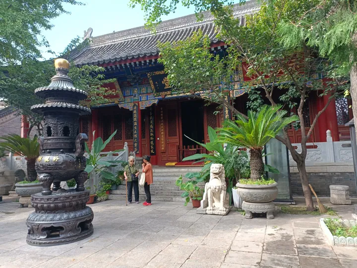 The courtyard of 卧龙寺: a hall with red doors, painted blue-and-gold brackets and a grey tiled roof on a raised stone terrace, a huge tiered bronze incense burner with a gilt finial in the left foreground, a stone lion on a pedestal at the right, potted cycads and two visitors in the middle.