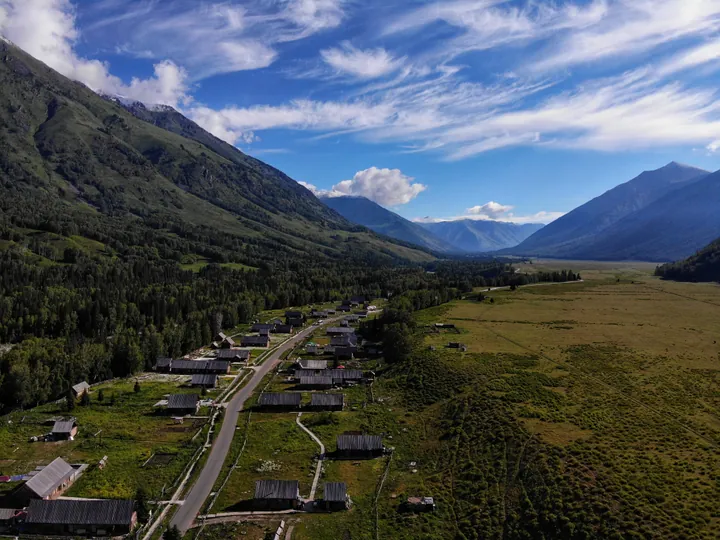 An elevated view down the 禾木 valley: a scattered village of dark log houses with plank roofs along a road, green meadows and a strip of crops at the right, dense spruce forest and steep slopes at the left, and snow-streaked mountains closing the valley under a deep blue sky streaked with cirrus.