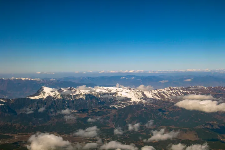 An aerial view of a snow-covered mountain massif: white ridges and dark rock running across the middle of the frame, wooded slopes and a valley with villages and fields below, cloud lying in the valleys and a clear deep blue sky above.
