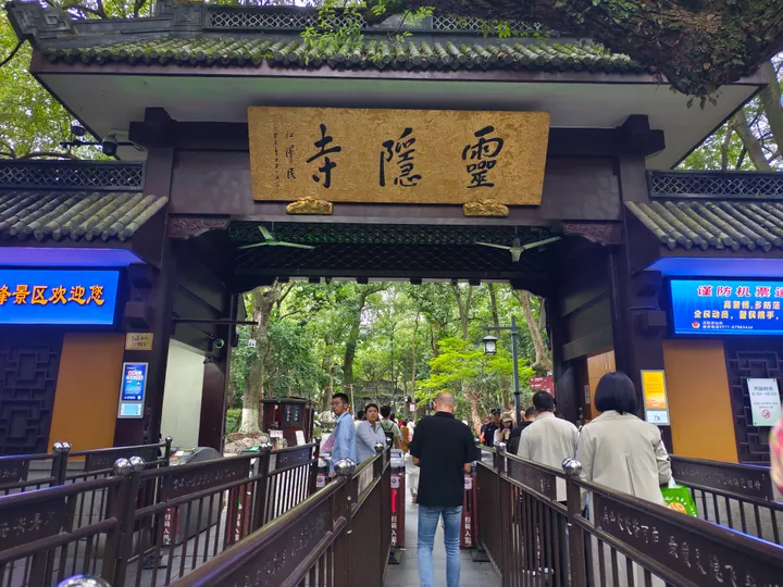 The east gate of the 灵隐飞来峰 scenic area: a dark timber gateway with grey tiled eaves and a gold plaque lettered 靈隱寺, ticket turnstiles and railings leading through it, visitors queuing, and electronic notice screens on both flanking walls beneath the trees.