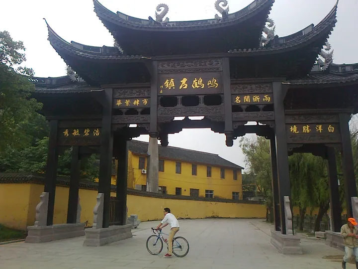 A large dark-timber paifang spanning a paved square: layered upturned eaves with dragon ridge figures, the central plaque 鳴鶴古鎮 and side plaques 市繁華 / 名聞浙雨 in gold, stone piers on carved bases, a cyclist passing beneath and a yellow building behind.