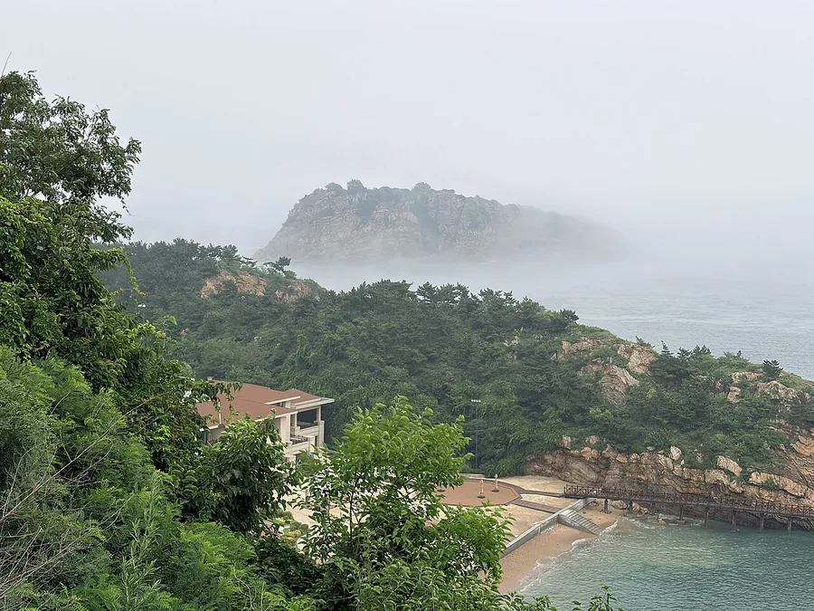 Bangchuidao Island seen through sea mist beyond the foreground shore in Dalian, Liaoning; 16 July 2026.
