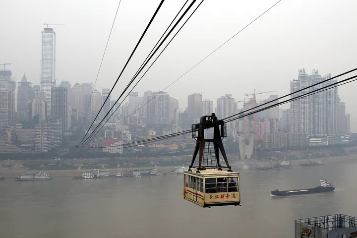 A cable car gondola suspended over a wide river, with a dense high-rise skyline in the hazy background
