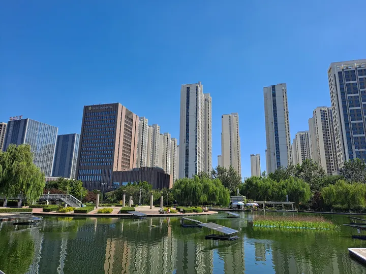 A city park lake with willow trees and modern high-rise buildings in the background, Lunan District
