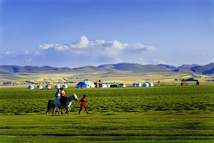 Riders on horseback beside yurts on a green grassland under a blue sky