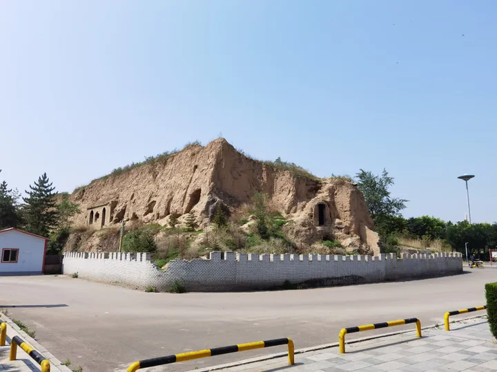 Eroded earthen city wall ruins with carved niches, enclosed by a low patterned wall