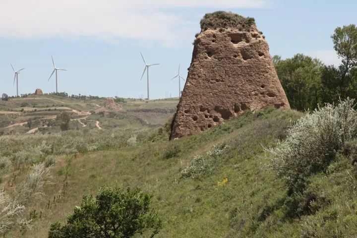 Eroded earthen Great Wall watchtower on a grassy hillside with wind turbines in the distance
