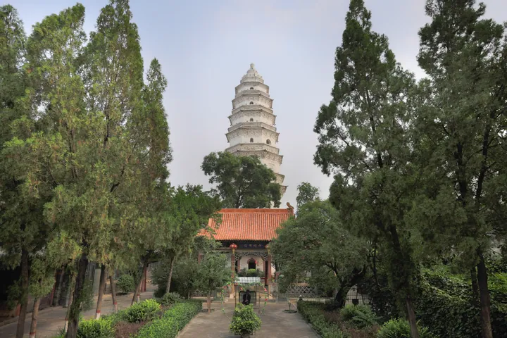A tall white pagoda seen through trees above a red-roofed temple hall, Taigu District
