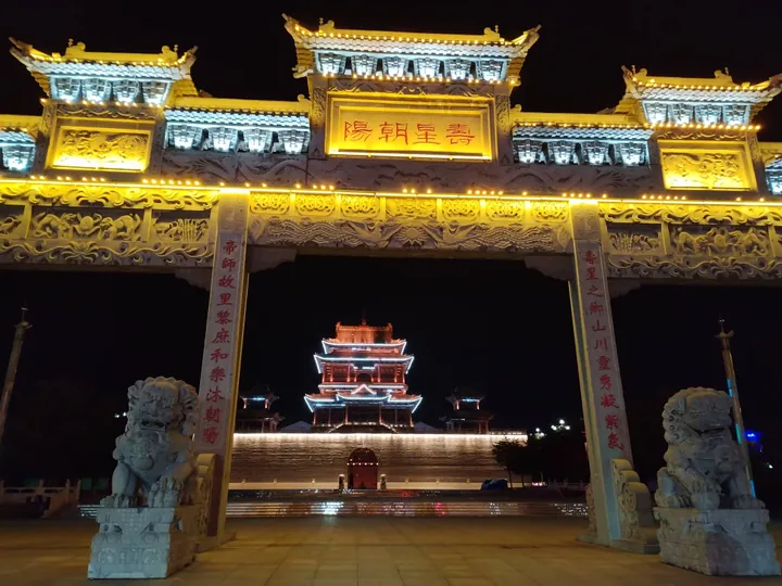 Ornate lit ceremonial archway with stone lions and a pagoda-style pavilion behind it at night