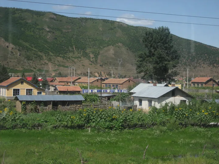 Rural village with red-roofed houses below wooded hills near Zhalantun