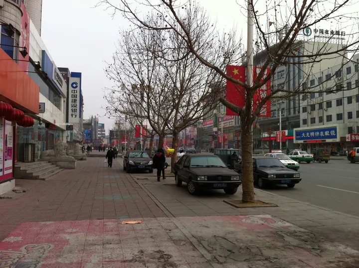 Tree-lined commercial street with shops and parked cars in Gaizhou