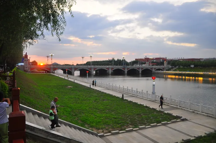 An arched bridge over a river at sunset with a lit riverside promenade in Yitong County