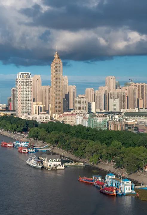 Skyline of Harbin seen across the river with tour boats moored under dramatic clouds