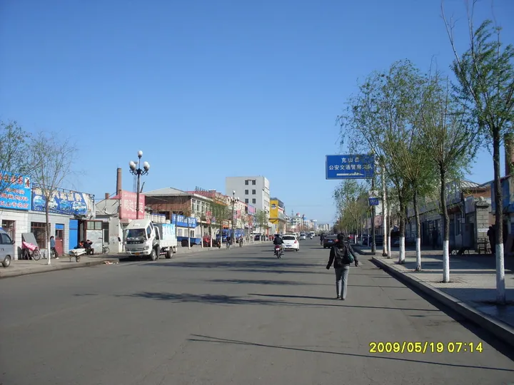 A street lined with shops and trees, with a road sign reading 'Keshan County', on a clear day