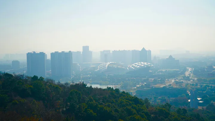 A hazy city skyline with high-rise buildings and a stadium seen from a hillside
