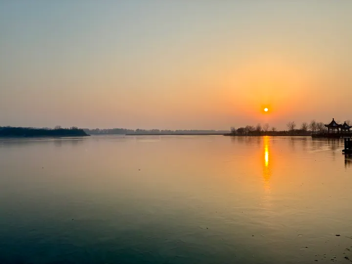 Sun setting over a calm wetland lake with pavilions in Donghai County