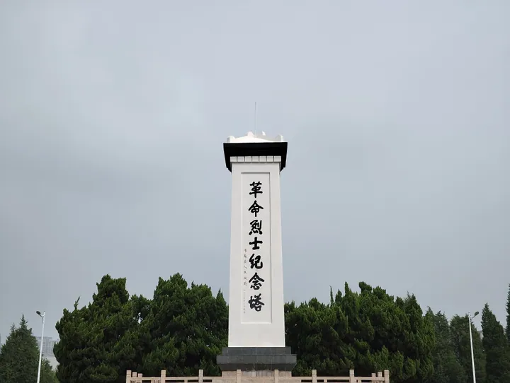 A tall white memorial column inscribed with Chinese characters, set against trees, in Guannan County