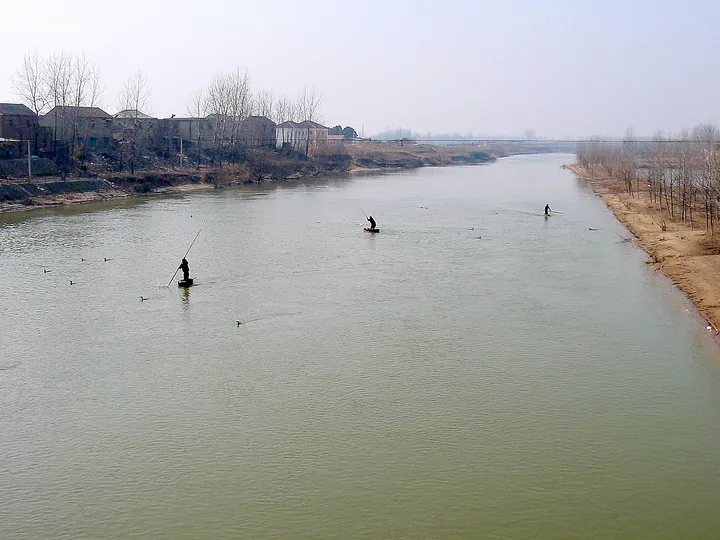 Two fishermen on small rafts on a wide river near village houses in Lianshui County