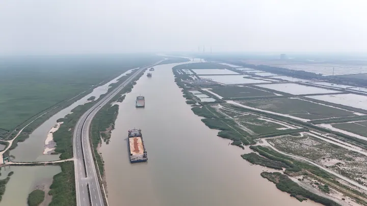 An aerial view of barges on a wide muddy river bordered by wetlands and farmland in Sheyang County