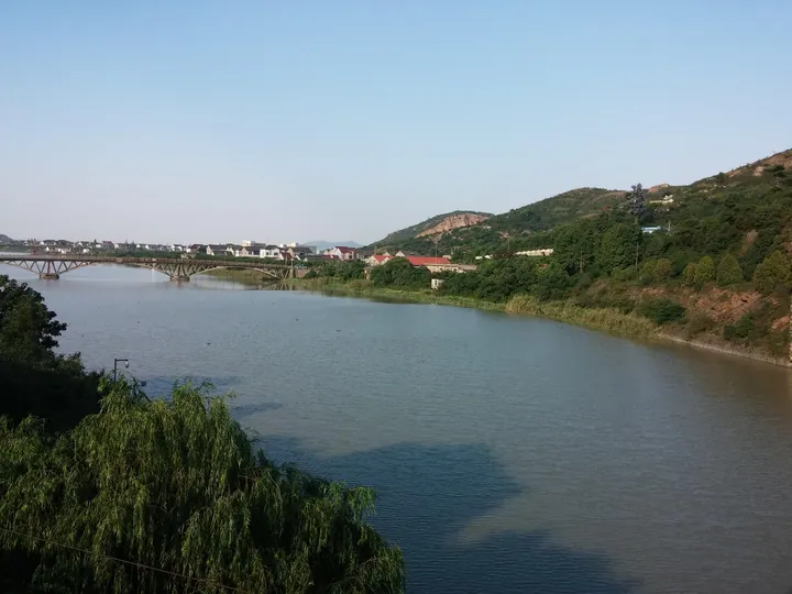 A calm river winding past a village, an arched bridge and green hills near Haiyan