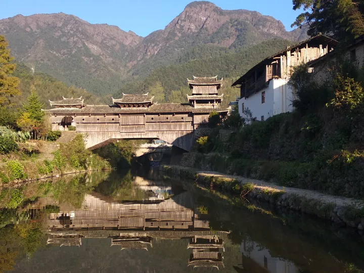 A traditional wooden covered bridge reflected in a river, backed by mountains