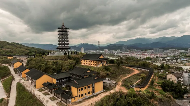 A tall traditional pagoda on a hillside temple complex with the city skyline behind under dramatic clouds