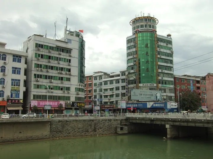 A riverside town street with a tall green office building reflected in the water in Xiaoxi, Pinghe County