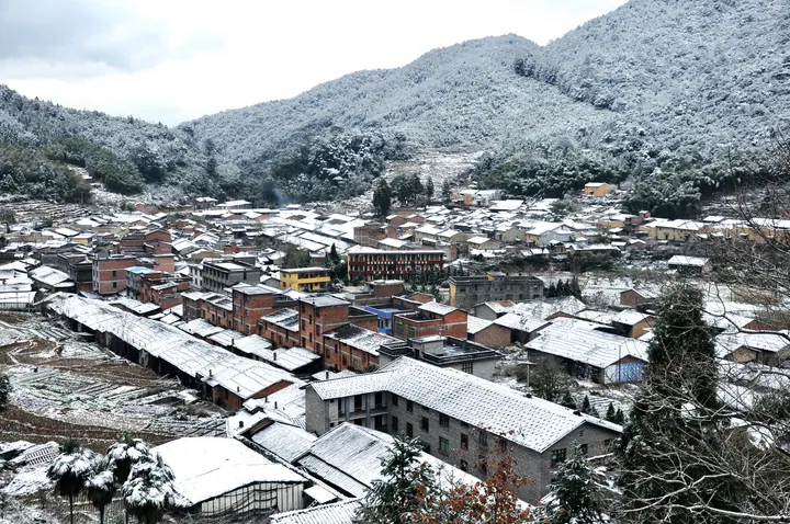 A hillside village covered in fresh snow surrounded by forested mountains in Gutian County