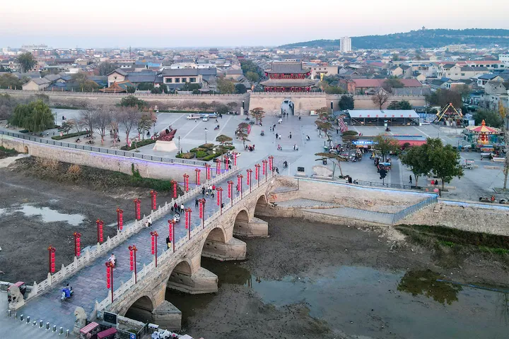 An aerial view of a stone bridge decorated with red lanterns leading to a city gate and plaza in Xun County
