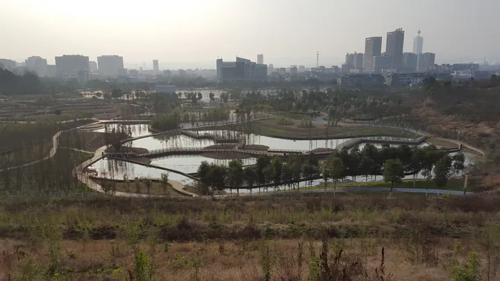 A landscaped lake park with a curved boardwalk and the city skyline in the background in Xiaoting District