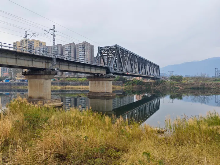 A steel railway bridge crossing a river with a city skyline and mountains behind