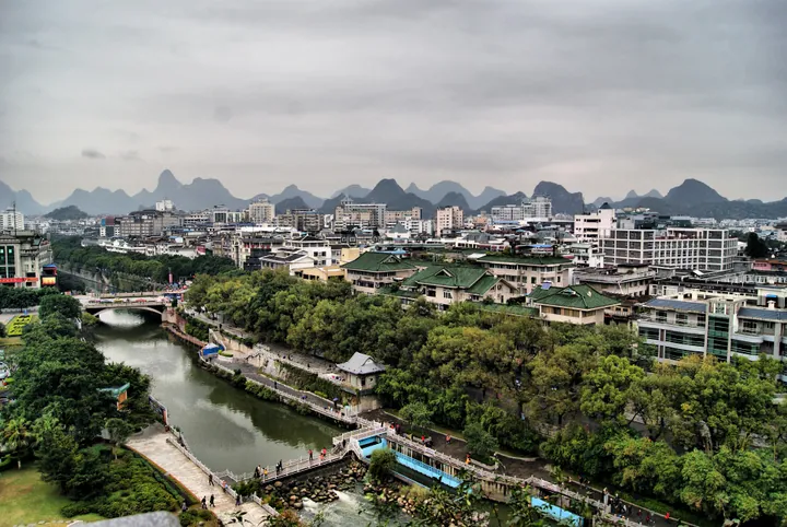 Green-roofed riverside pavilions in front of a hazy karst mountain skyline