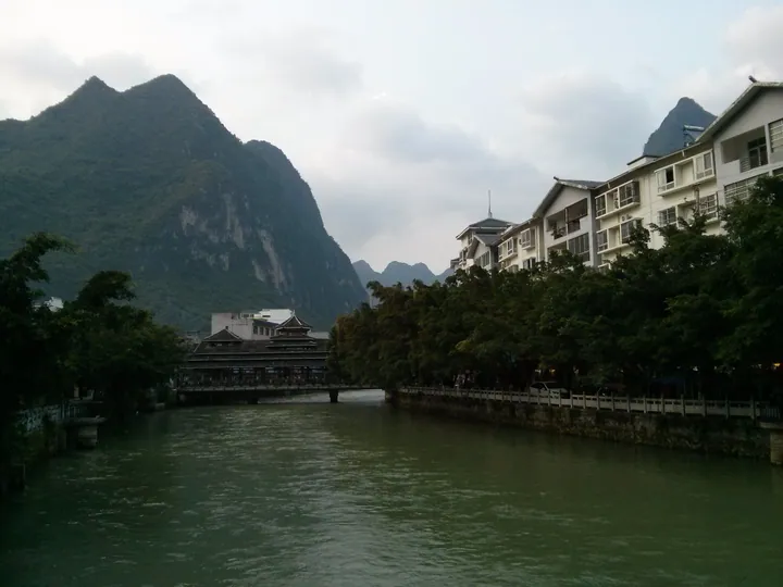 A river with a bridge, boats and riverside buildings below green karst hills in Lingyun County