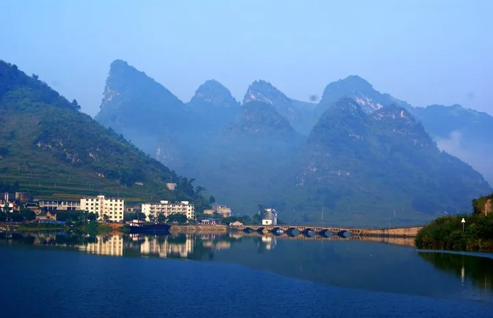 A lake reflecting karst mountains with a stone bridge and riverside buildings in Bama Yao Autonomous County