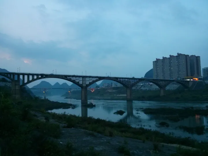 Two arched bridges crossing a river at dusk, with hills and a high-rise building behind