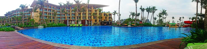 A large curved resort swimming pool lined with palm trees and hotel buildings in Haitang Bay, Sanya