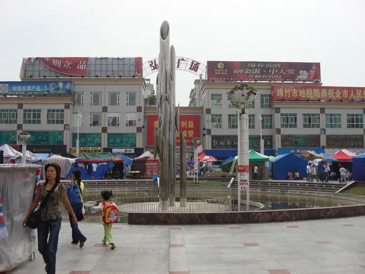 Public square with a metal sculpture, market tents and pedestrians in Mianzhu