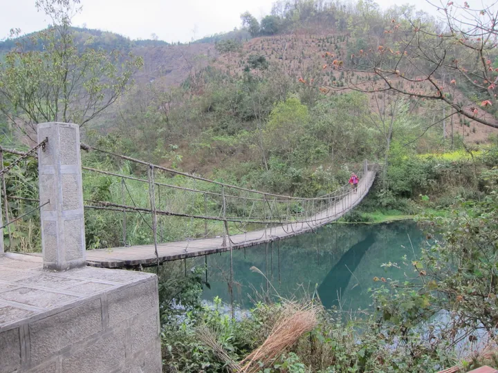 Rope suspension bridge crossing a turquoise stream in green hills