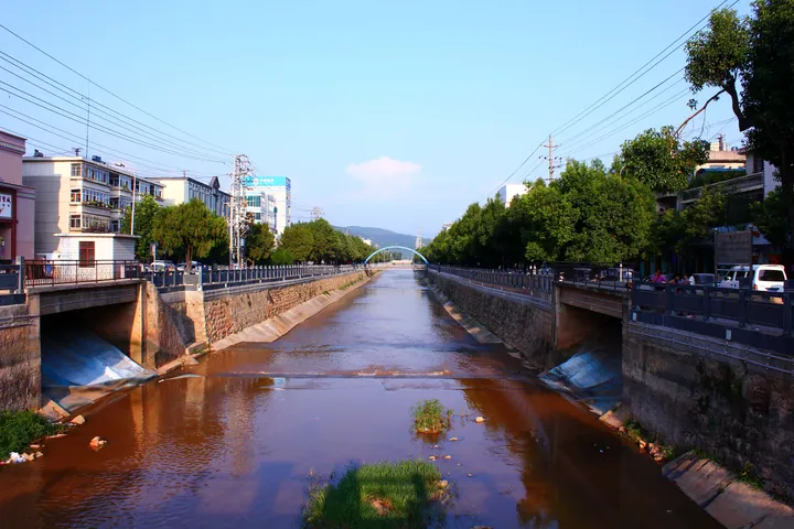 A canalised river lined with buildings and trees under a blue sky in Eshan County