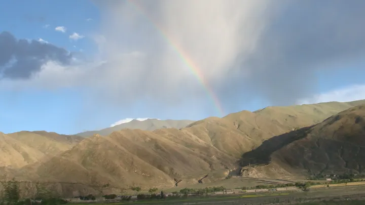 A rainbow arching over dry brown mountains with a small village below, near Lhasa
