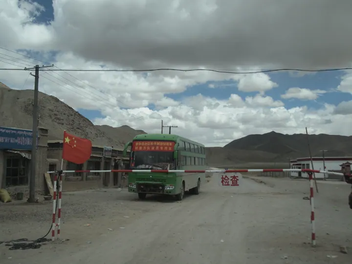 A roadside checkpoint with a barrier gate and a bus, houses and mountains behind, in Gar County, Tibet.