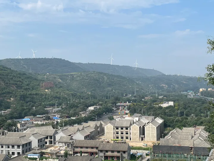 Traditional-roofed buildings on a hillside with wind turbines on distant ridges