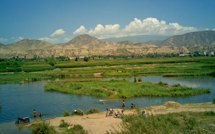 People and bicycles by a river with mountains and a village in the background, Lintao County