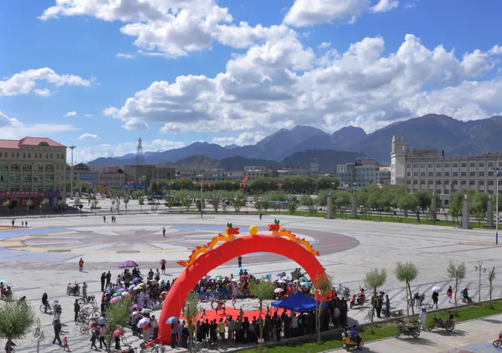 People gathered under a red dragon arch on a city square with mountains behind, Delingha