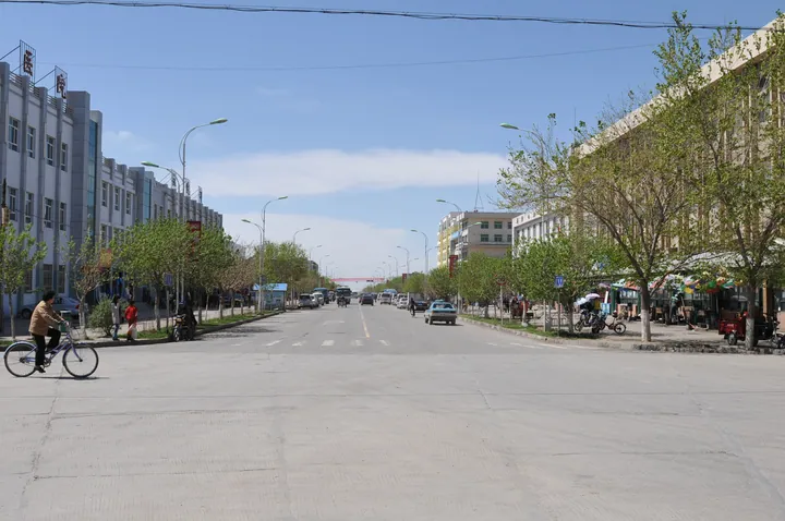 A wide town street with buildings, trees and a cyclist under a blue sky