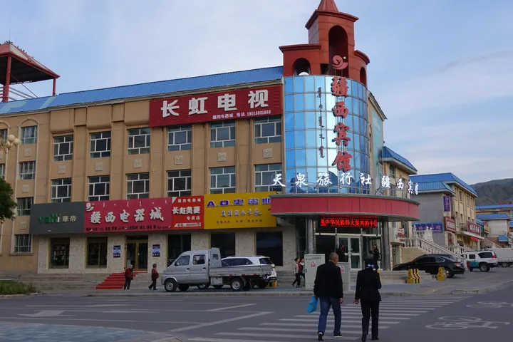 A street corner with a hotel building, parked vehicles and pedestrians