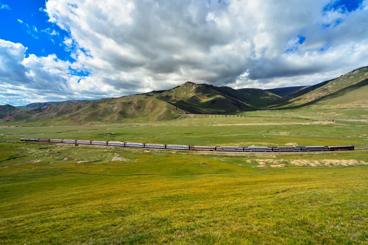 A passenger train crossing green grassland with mountains behind, in Hejing County