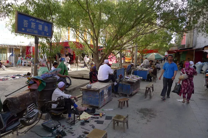 A tree-shaded market street with vendors, shoe-repair stalls and pedestrians, street sign reading Wusitang Lu